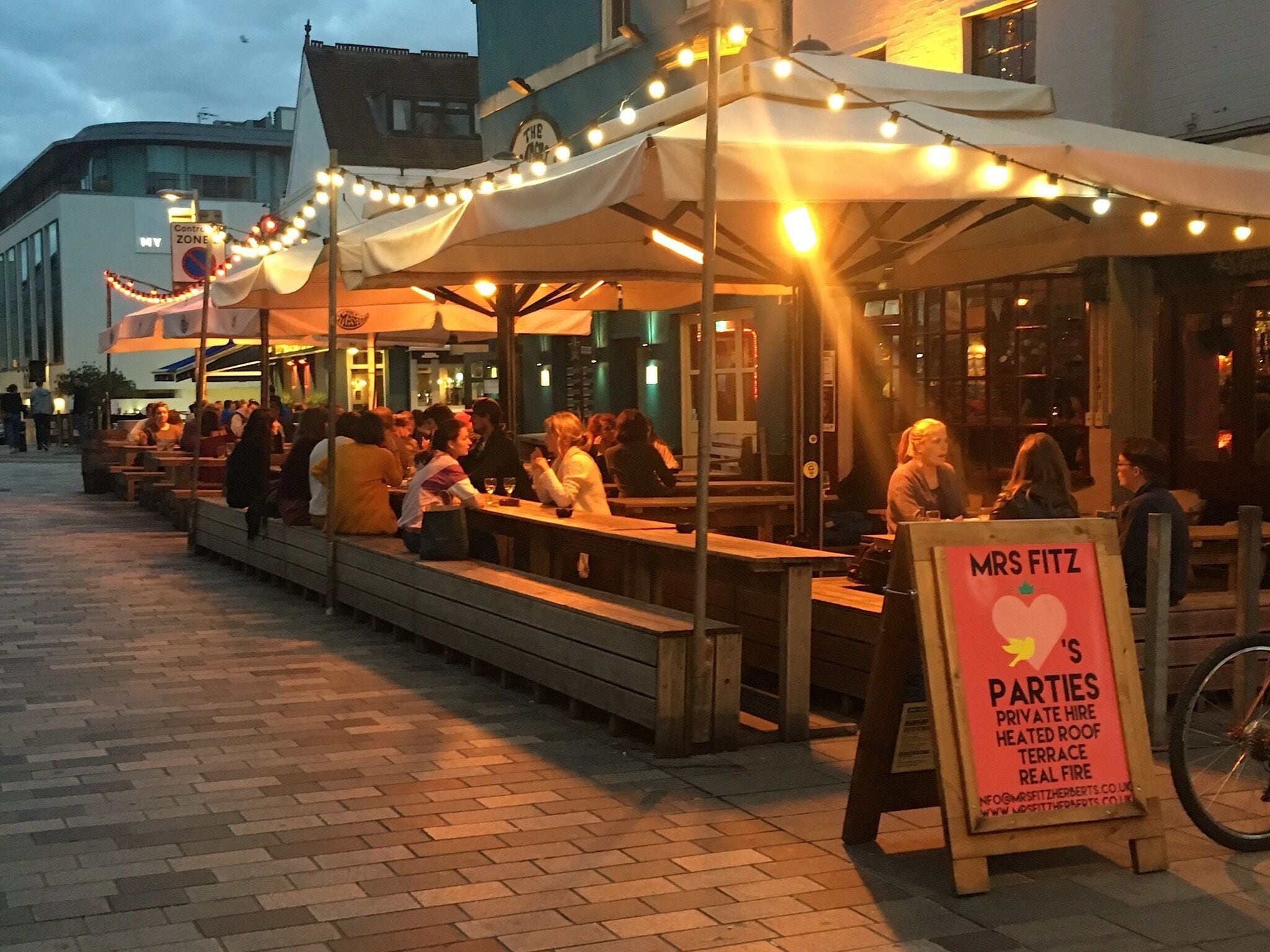 Een terras met tafels en banken onder lichtslingers, een bord met 'Mrs. Fitz's Parties' en een fiets in de buurt.