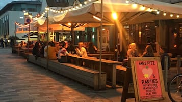 Een terras met tafels en banken onder lichtslingers, een bord met 'Mrs. Fitz's Parties' en een fiets in de buurt.