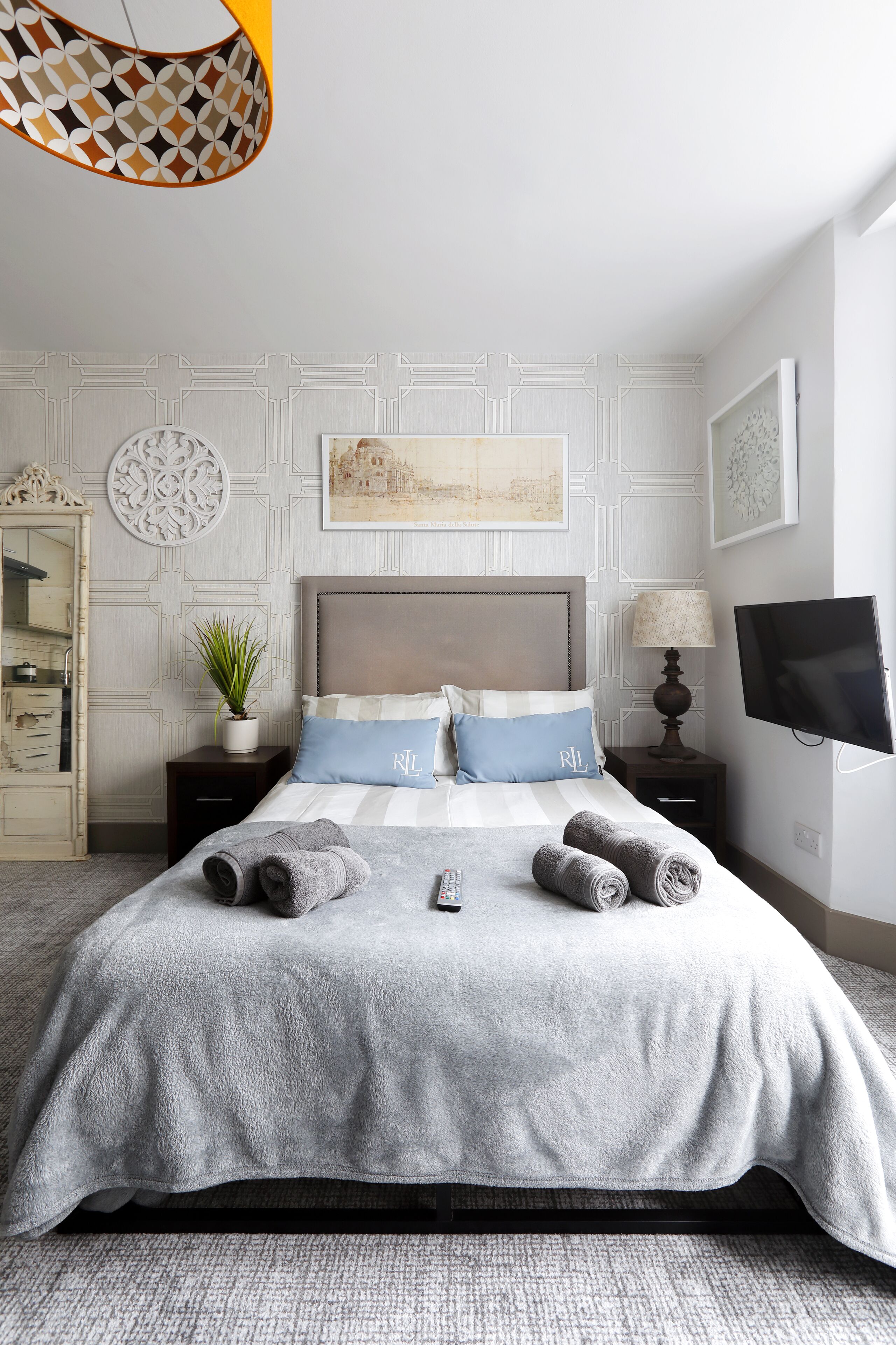 A neatly arranged bedroom with a bed, bedside tables, a lamp, a TV, and a decorative wall piece.