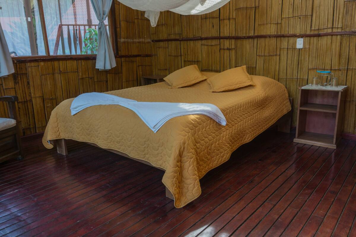 Una cama con cubrecama acolchado, una mesita de noche y una silla en una habitación con suelo de madera y paredes de bambú.