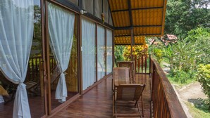 Una terraza de madera con un espacio de estar techado, puertas de vidrio grandes con cortinas y vistas a vegetación exuberante.