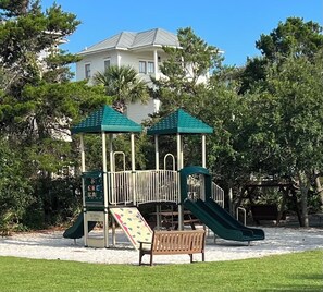 Children can play on the playground structure with multiple slides, surrounded by a grassy area and a nearby bench.