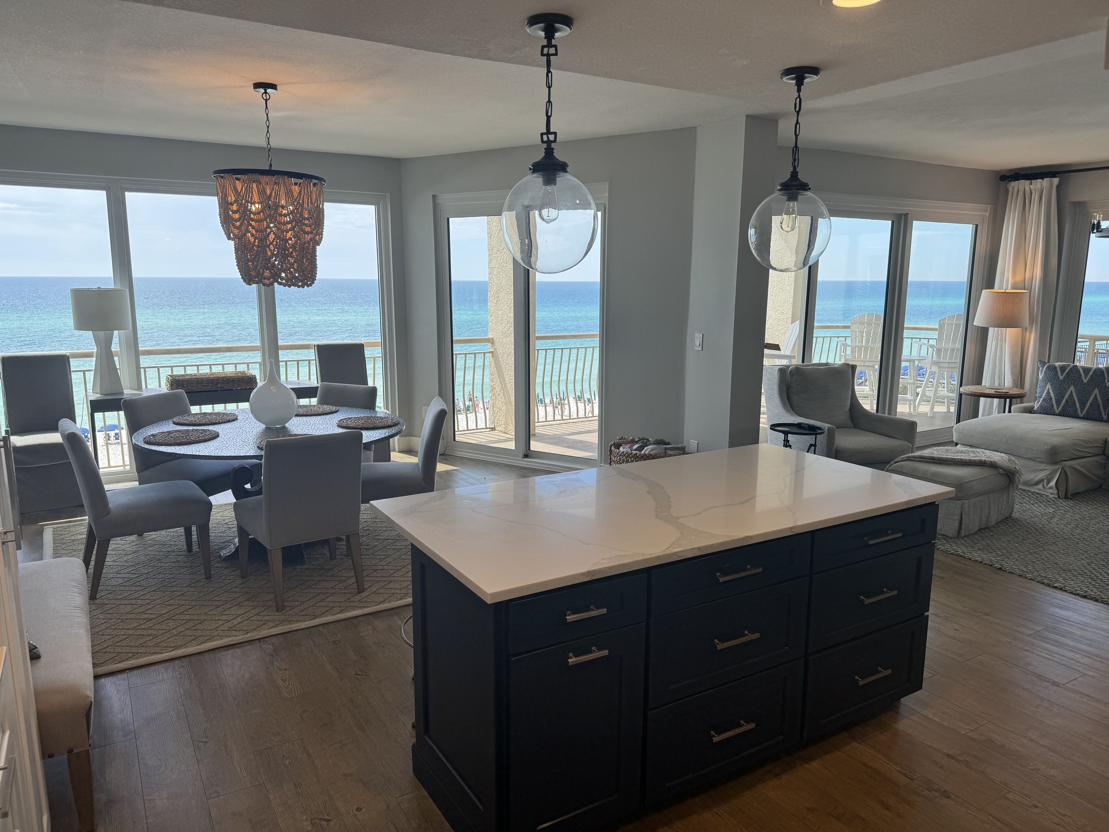 A spacious living area with a large island, dining table, and chairs, overlooking a balcony with a view of the ocean.