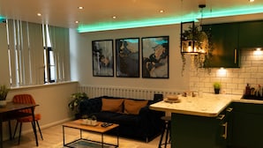 A modern living room with a dark blue sofa, a wooden coffee table, and a kitchen area with green cabinets and a white countertop. - The Opulence (Bradford)