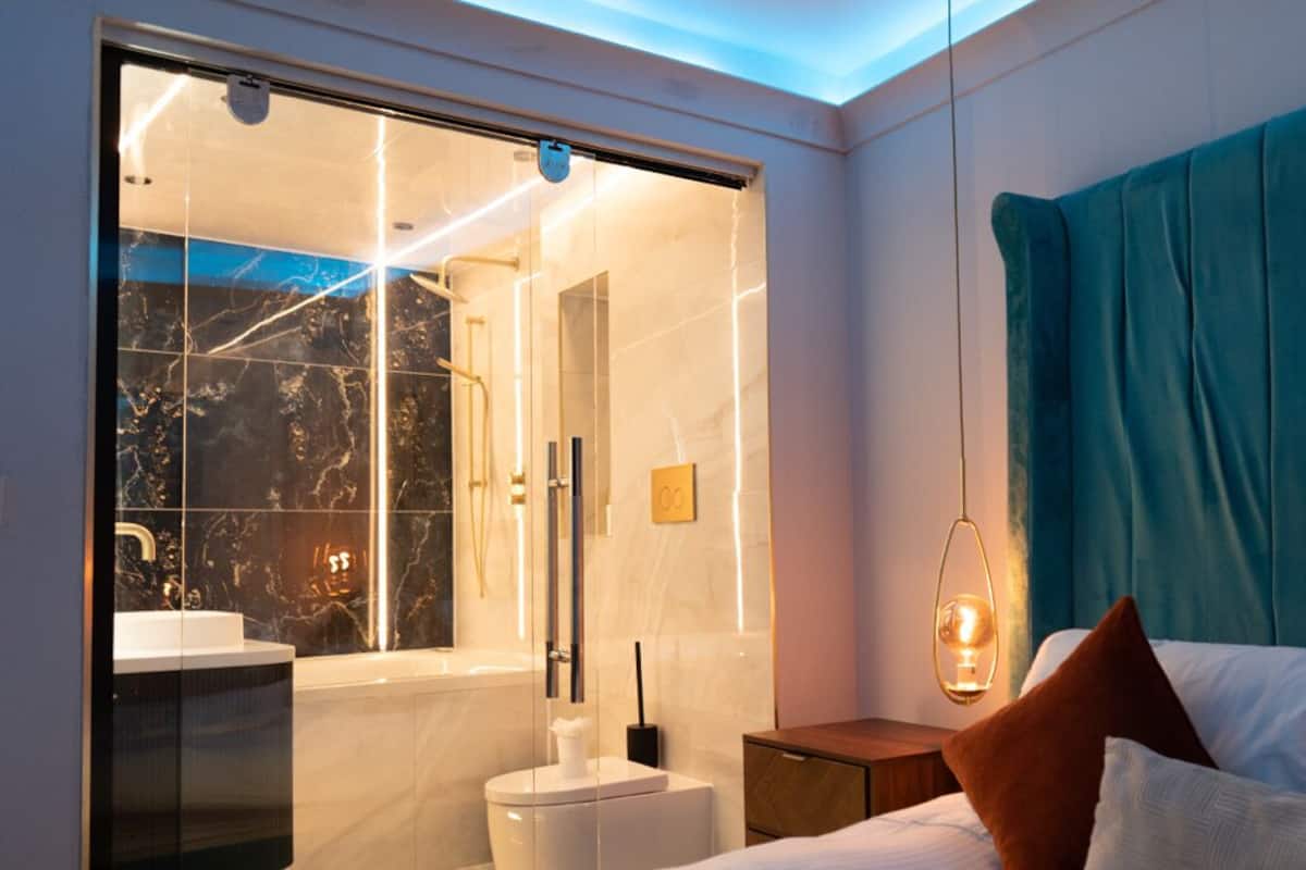 A modern hotel room with a glass shower enclosure, a bed with teal curtains, and a bedside table with a lamp.