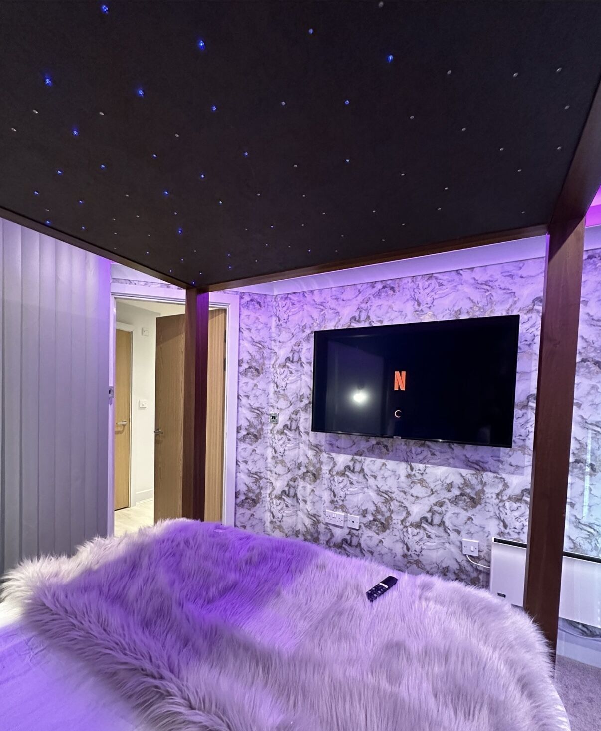 Un dormitorio moderno con una televisión de pantalla plana grande montada en una pared de mármol, una cama púrpura esponjosa y un techo con diseño de estrellas.