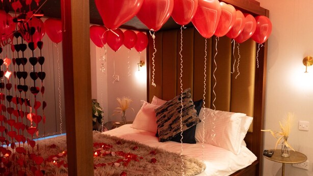 A bedroom with a bed, pillows, a bedside table with a vase, and red balloons. - The Opulence (Bradford)