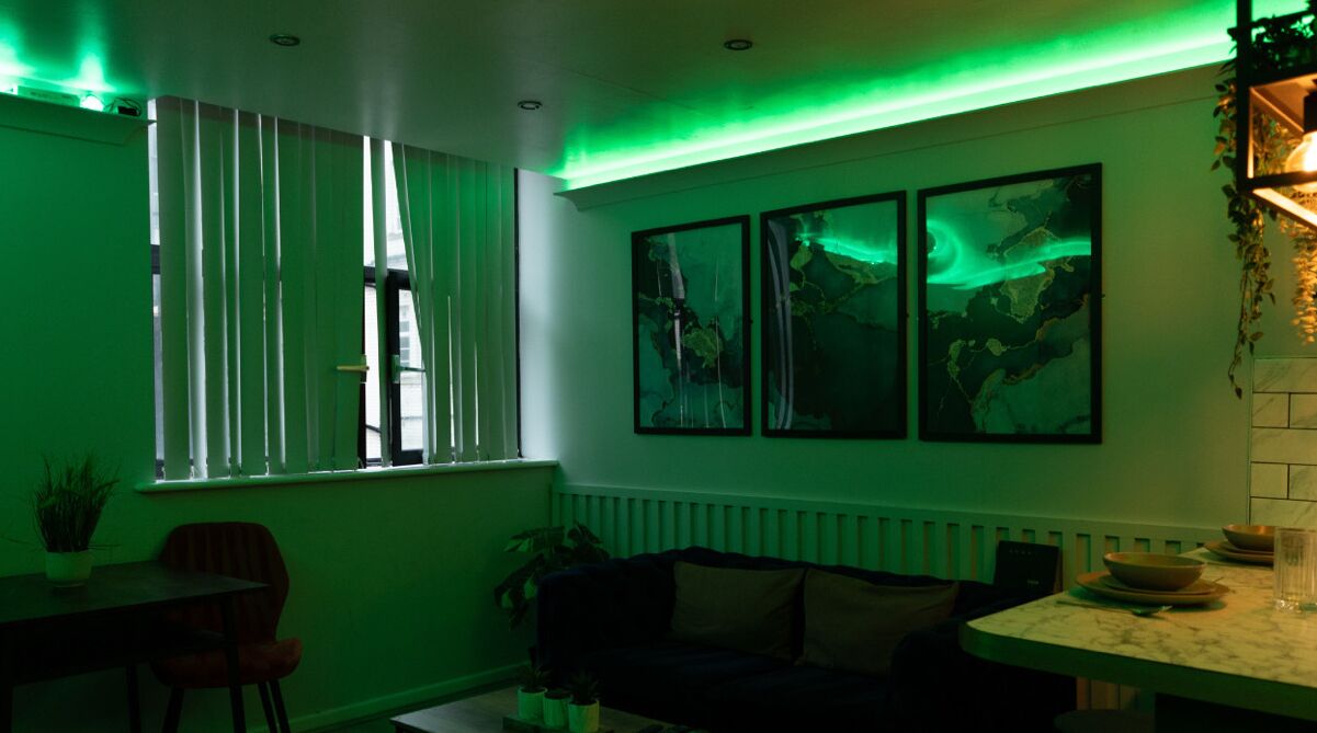 A room with a dining table, a sofa, and a window with vertical blinds. There are three framed pictures on the wall.