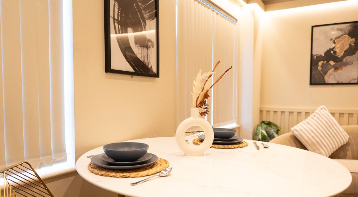 Un coin repas avec une table ronde, des bols noirs, un vase blanc contenant des plantes séchées et, au mur, un tableau abstrait encadré.
