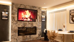 A modern living room with a fireplace, a flat-screen TV mounted on a marble wall, a dining area with a round table and chairs, and a large window with blinds. - The Opulence (Bradford)