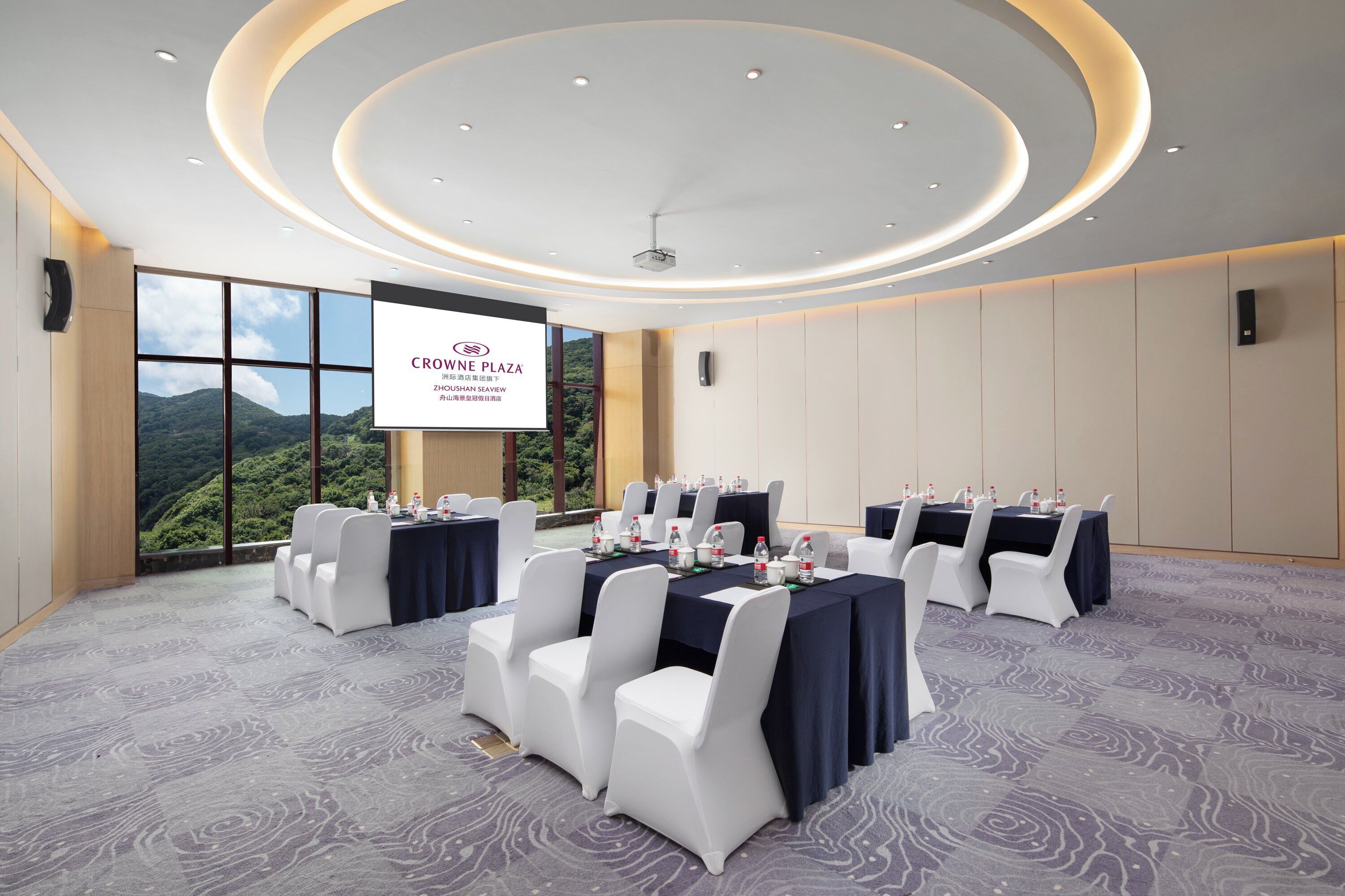 A conference room with tables set for a meeting, a large screen displaying text, and a view of a mountainous landscape through large windows.