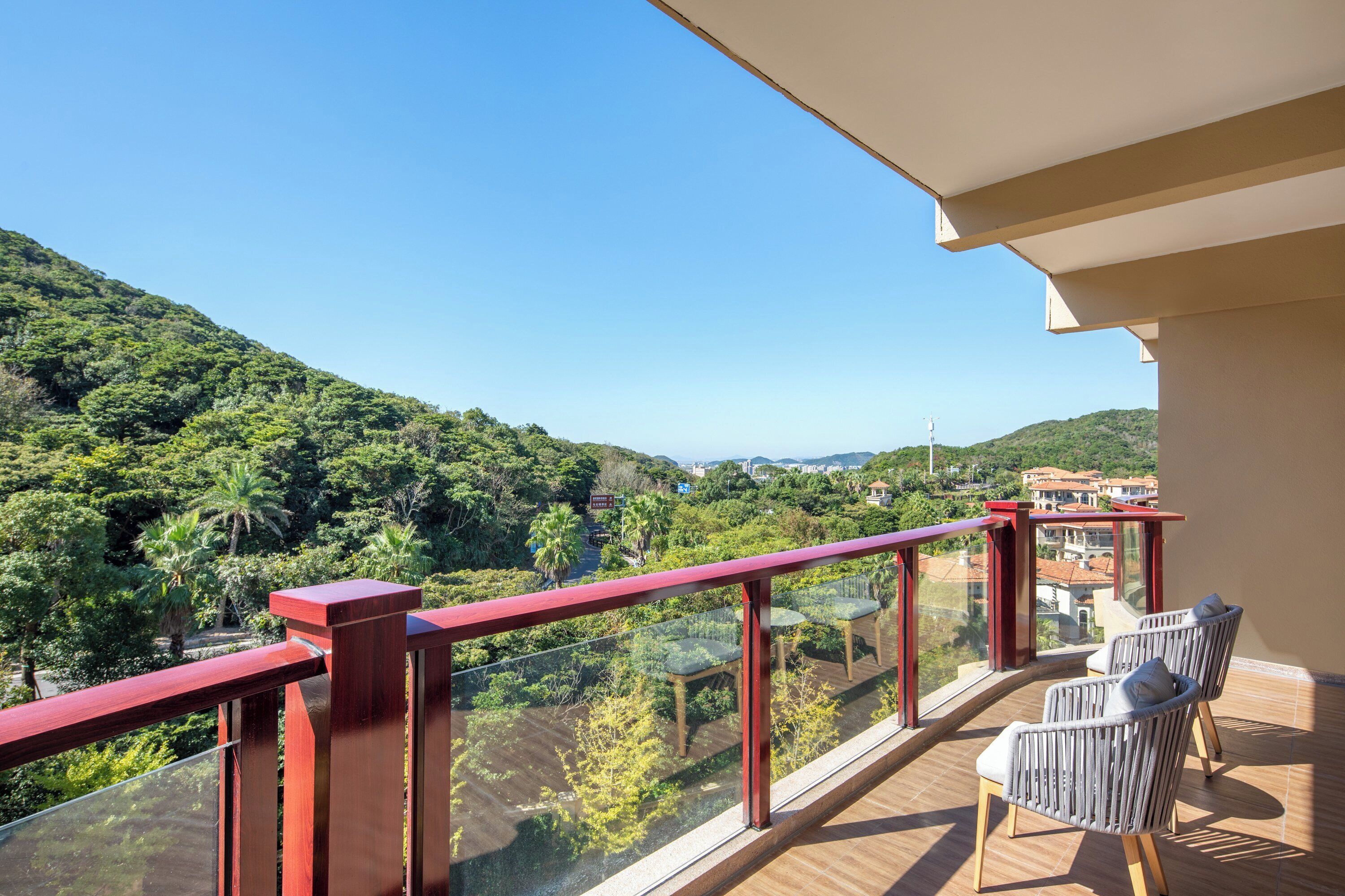 A balcony with a glass railing, two wicker chairs, and a view of a green valley with trees and a distant cityscape.