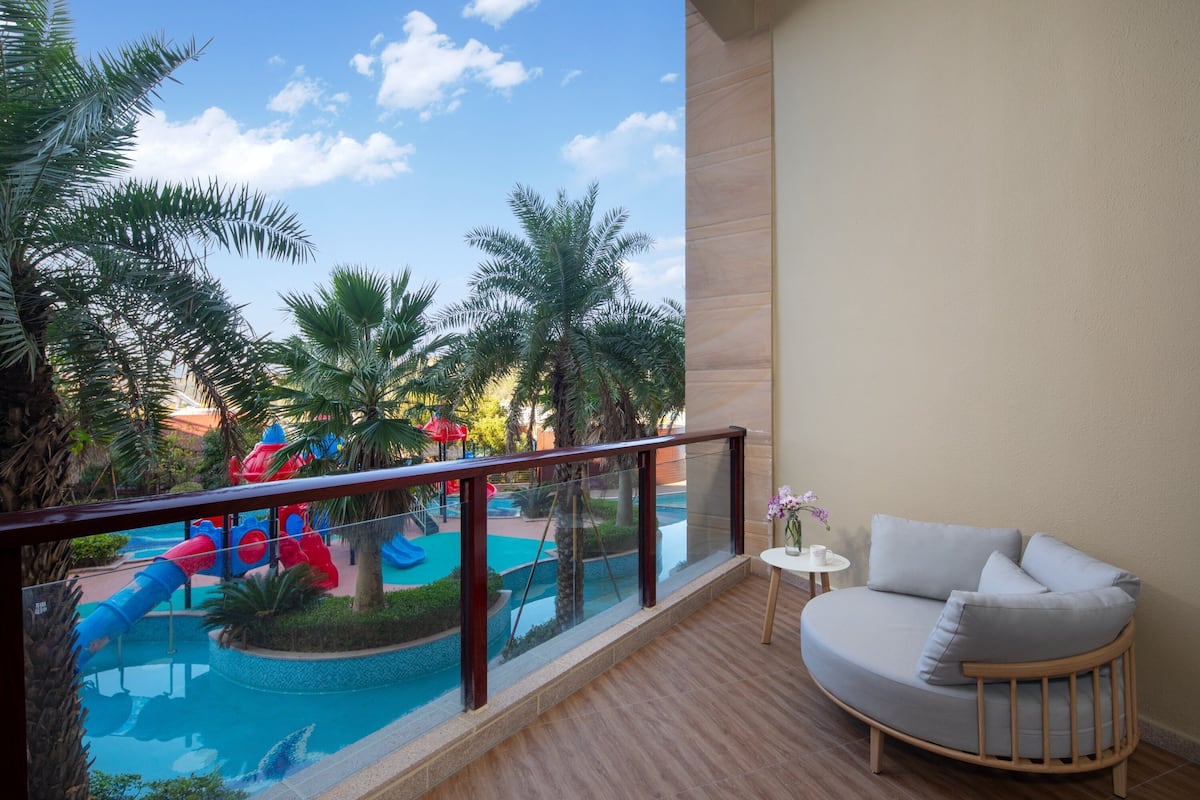Uma varanda com vista para uma piscina e toboáguas, um sofá branco com almofadas, e uma pequena mesa com um vaso de flores.