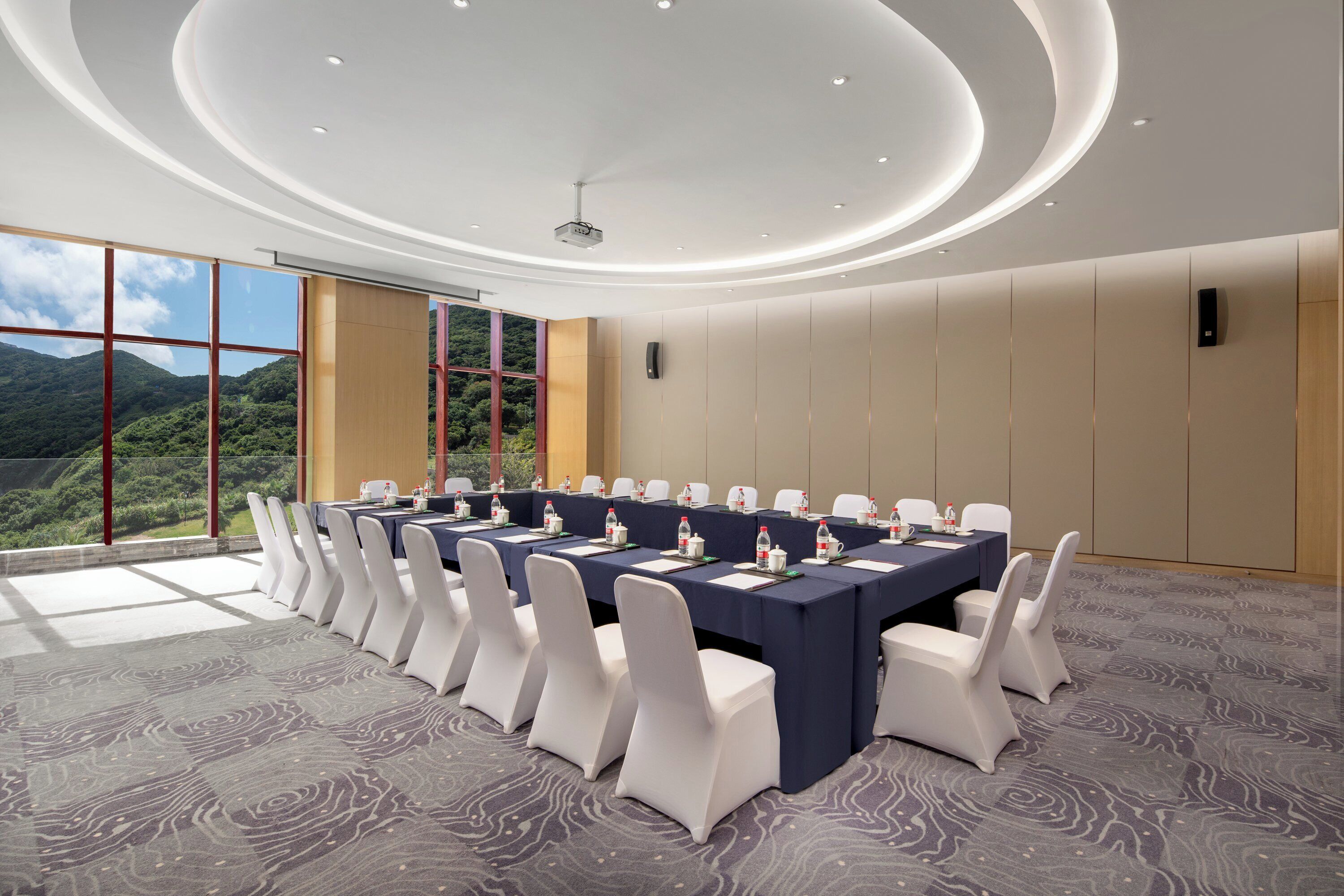 A conference room with a long table, white chairs, and a view of a mountainous landscape through large windows.