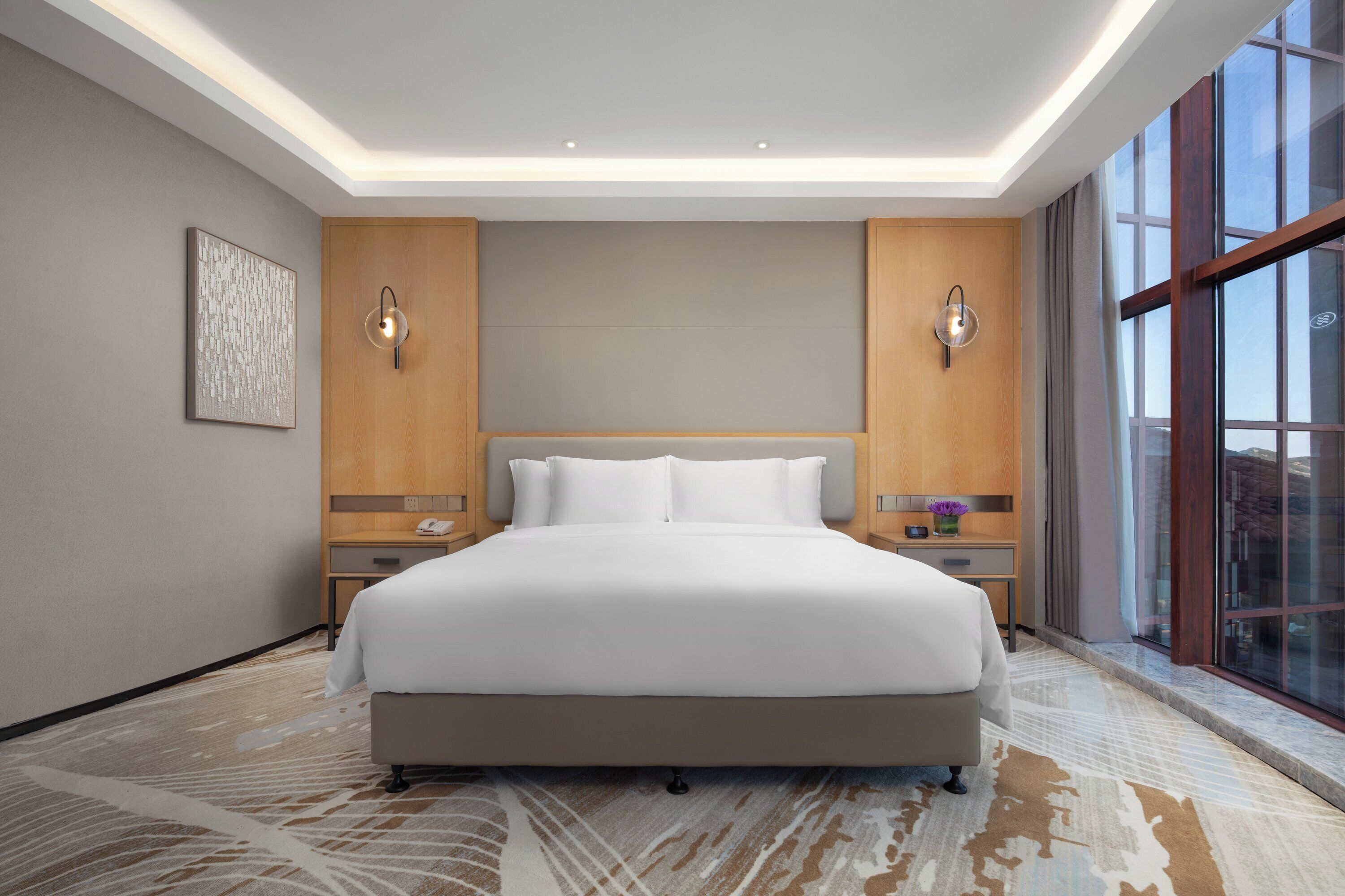 A hotel room with a large bed, two bedside tables, a nightstand, a wall-mounted lamp, and a large window.