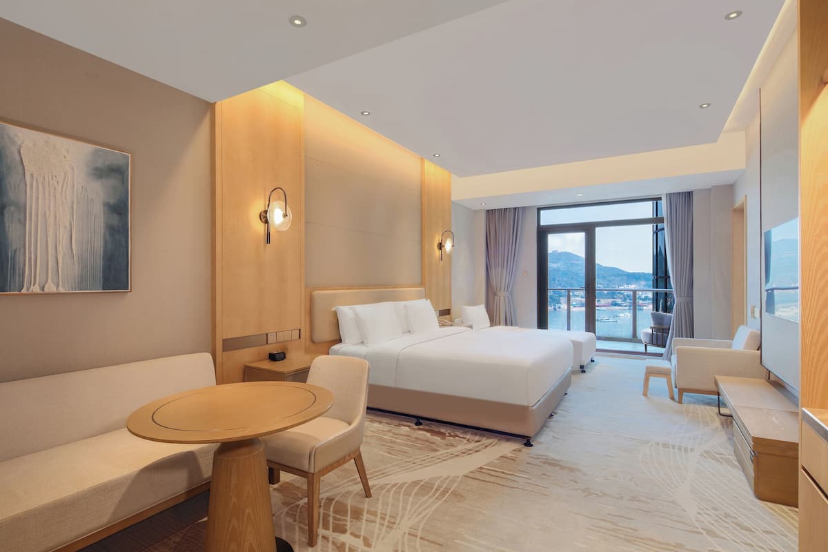A modern hotel room with a large bed, a sofa, a dining table, and a view of the city.