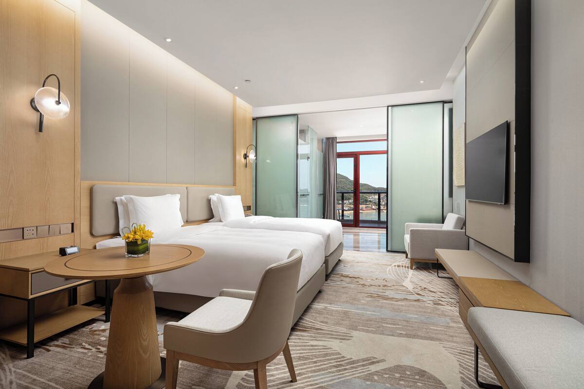 A modern hotel room with a large bed, a desk, a chair, a TV, and a view of the outdoors.