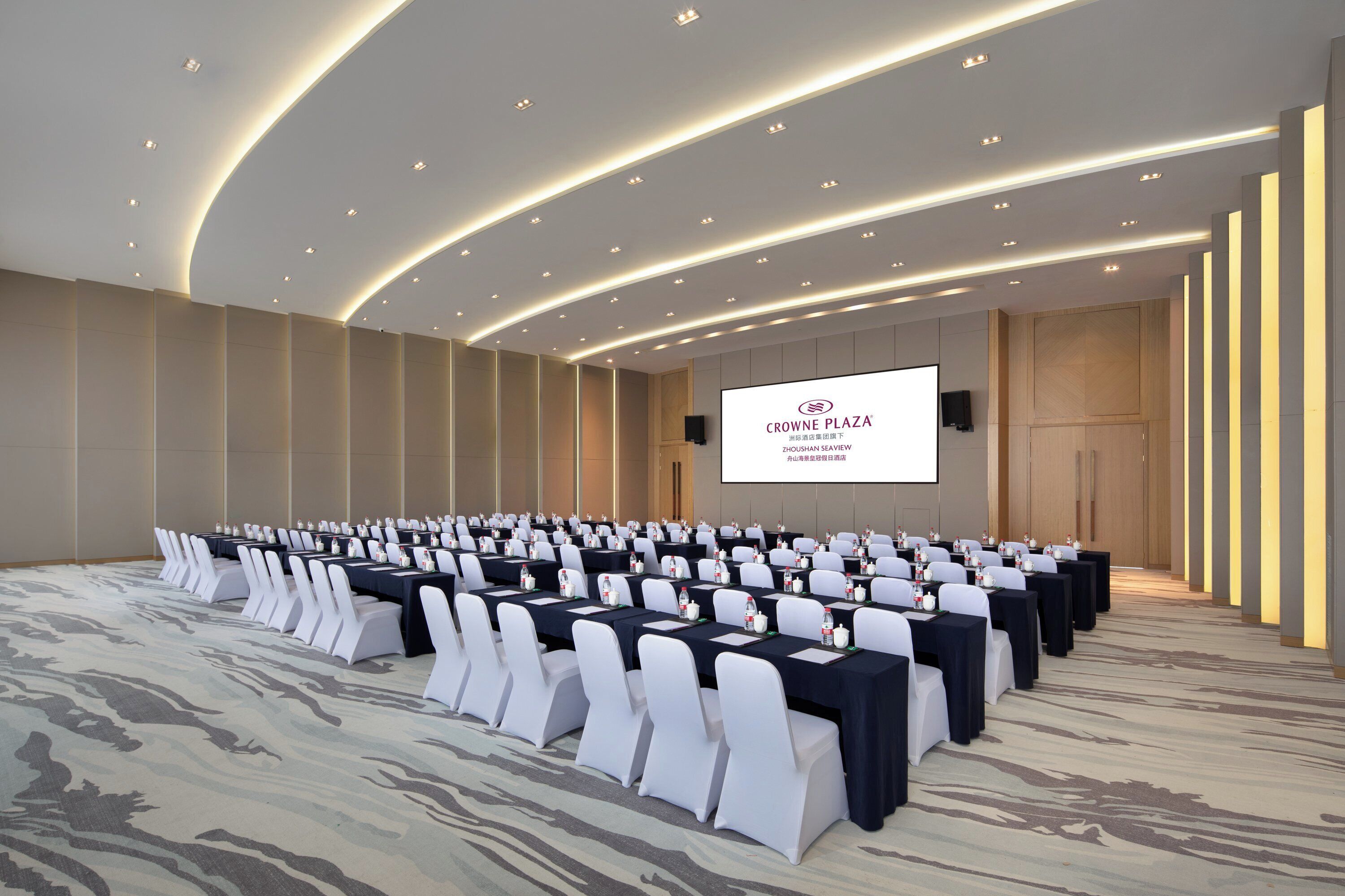A conference room with rows of tables and chairs, a large screen, and a modern ceiling design.