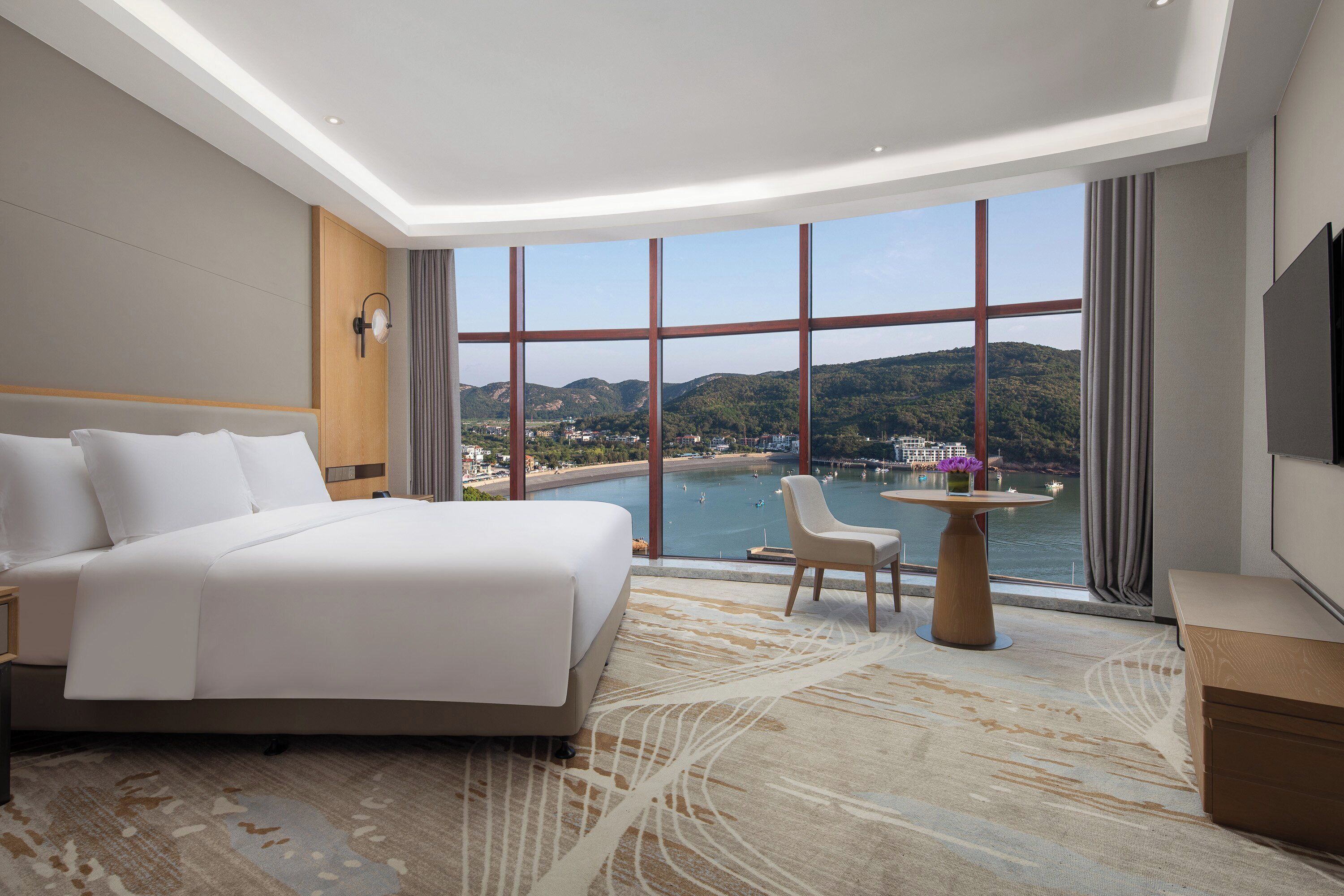 Ein Hotelzimmer mit einem großen Bett, einer Sitzecke und Blick auf ein Gewässer und Hügel.