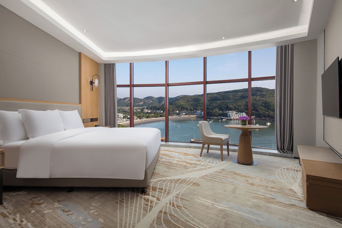 Une chambre d’hôtel dotée d’un grand lit, d’un coin salon et offrant une vue sur un plan d’eau et des collines.