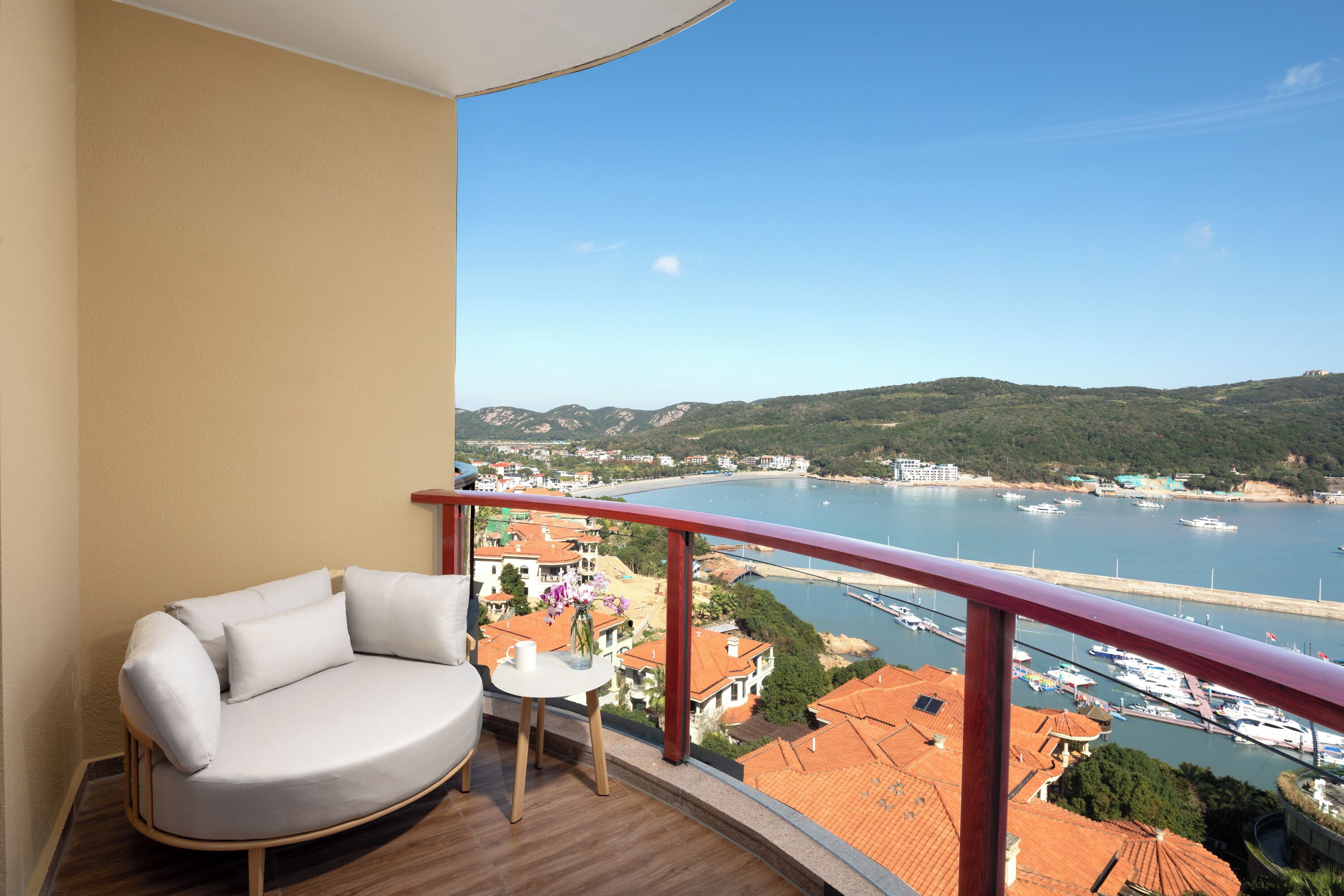 A balcony with a white sofa and a small table, overlooking a marina with boats and buildings.