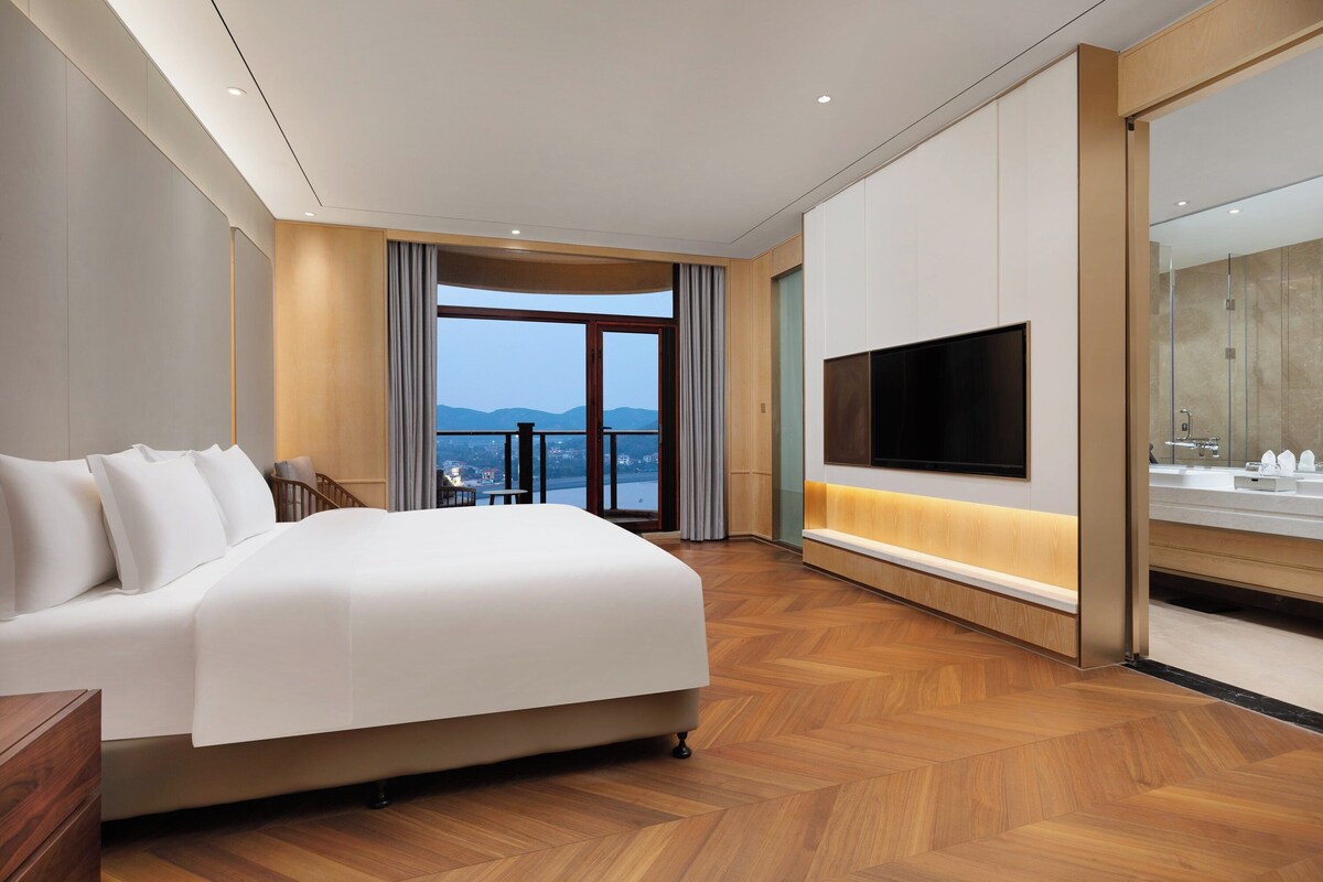 A hotel room with a large bed, a TV, a bathroom with a mirror, and a view of the city.