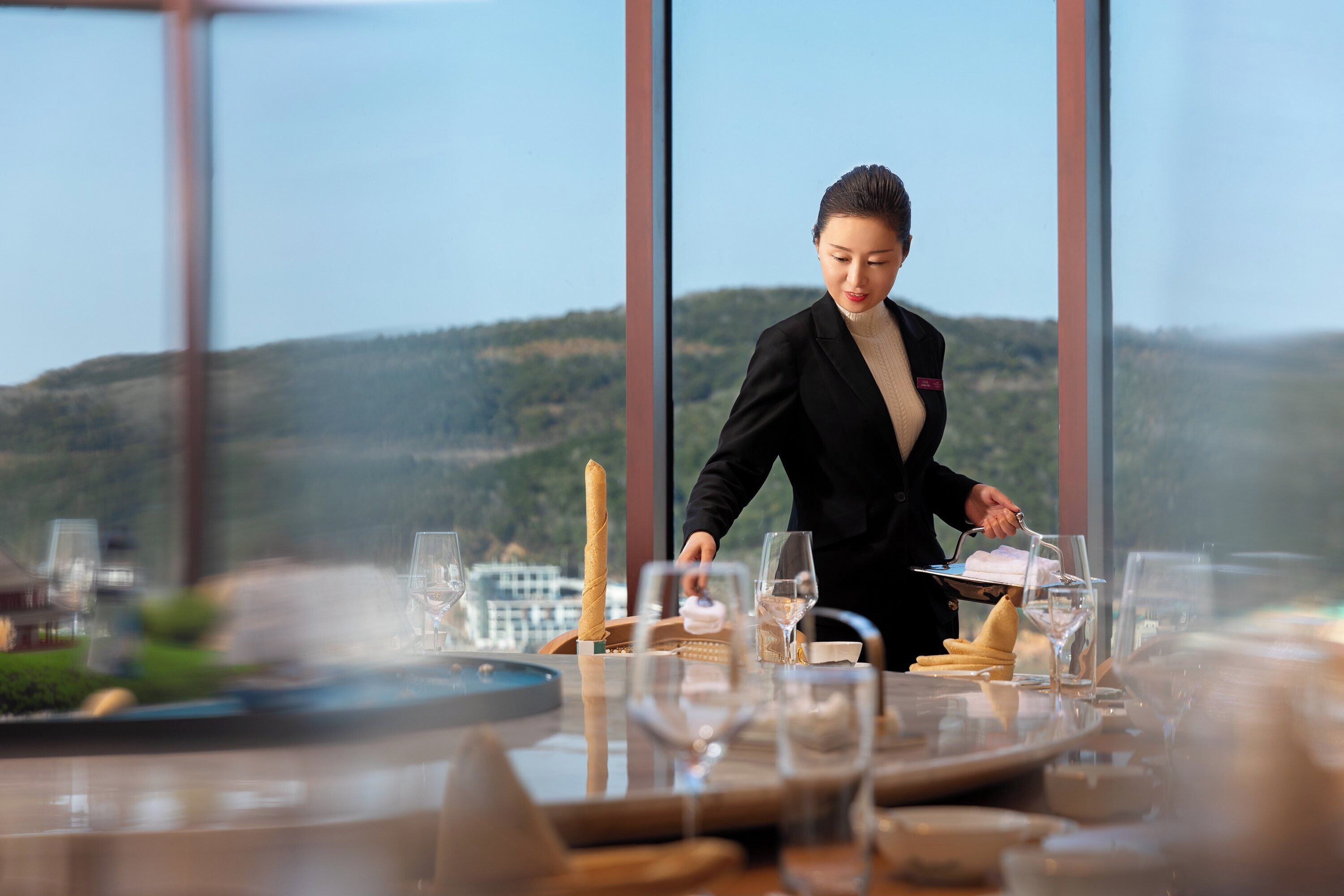 A person serving food at a table with a view of a hilly landscape through large windows.