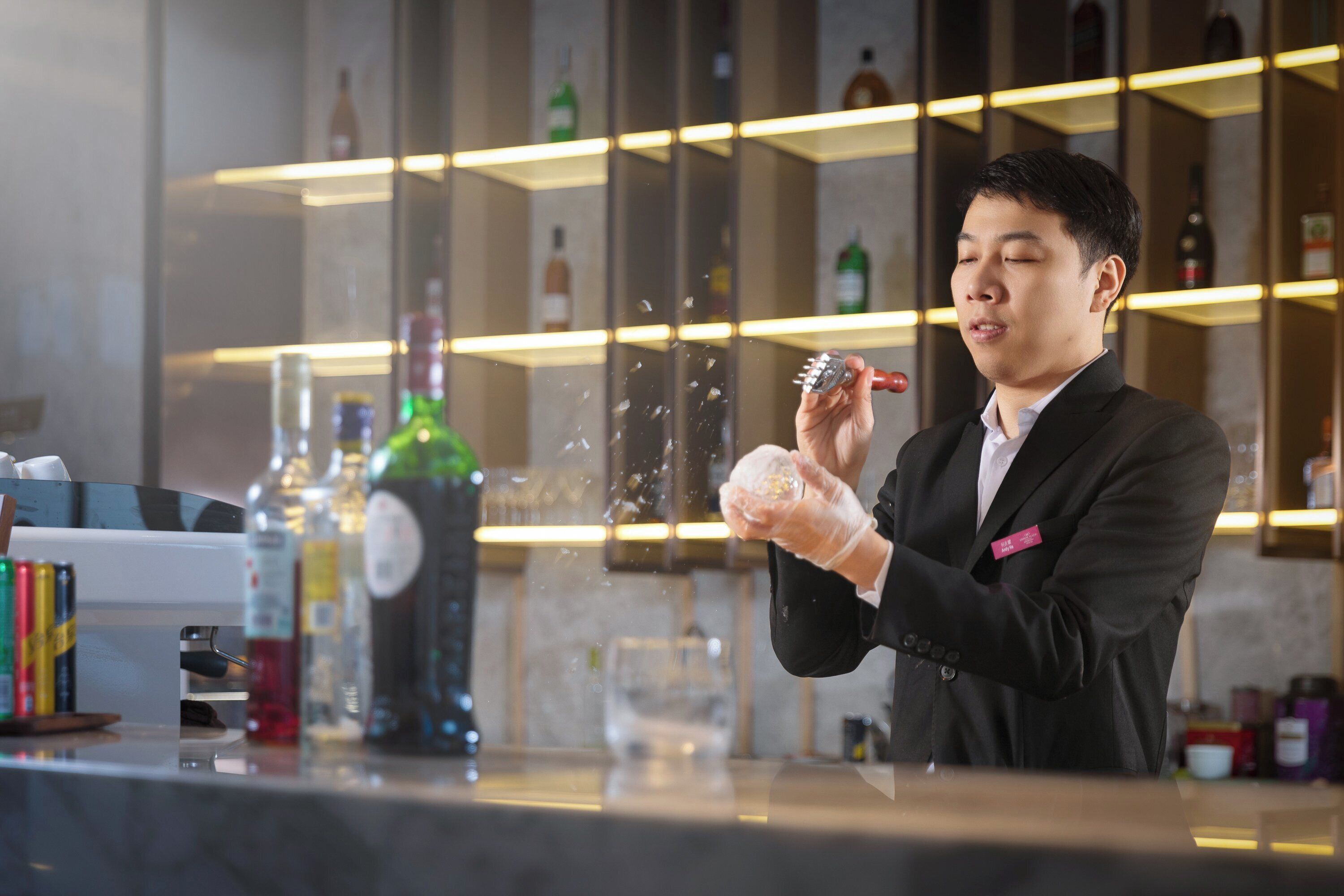 A person in a black suit holding a glass with a drink, standing in a bar area with shelves of bottles and a counter with various items.