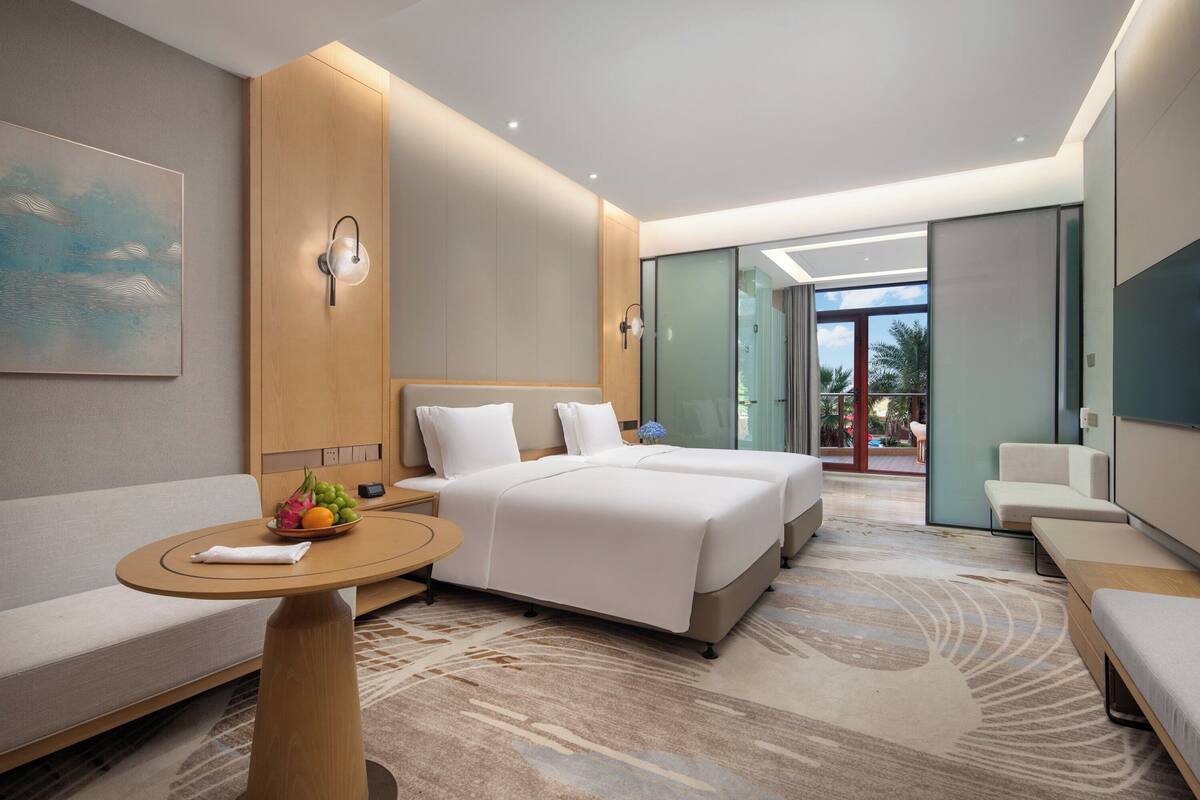 A modern hotel room with a large bed, a sofa, a small table with fruit, and a view of the outdoors.