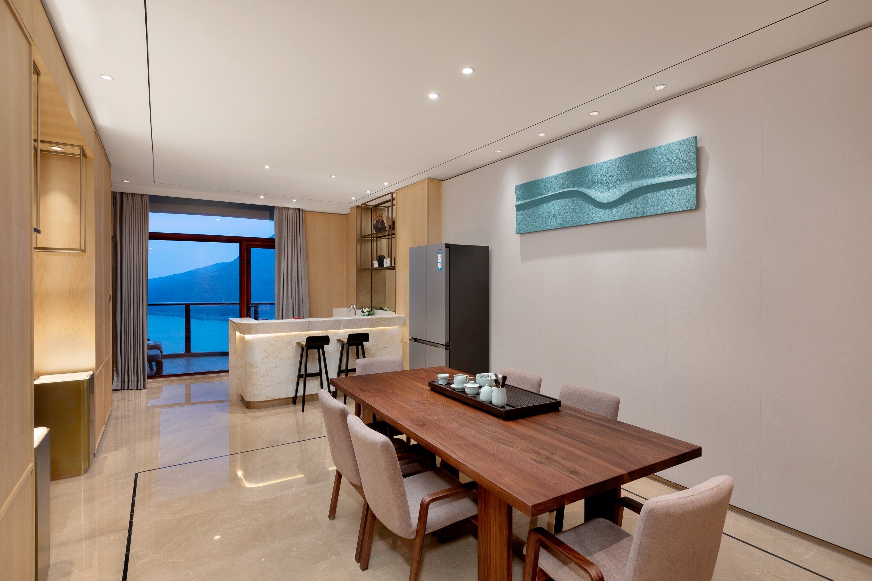Een moderne hotelkamer met een houten eettafel, gestoffeerde stoelen, een keukenhoek en een groot raam met uitzicht op zee.