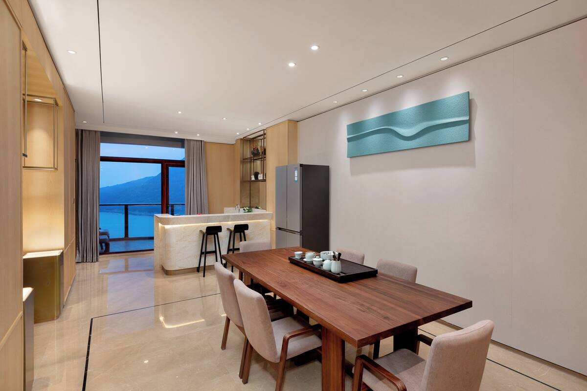 A modern hotel room with a wooden dining table, cushioned chairs, a kitchen area, and a large window offering a view of the sea.