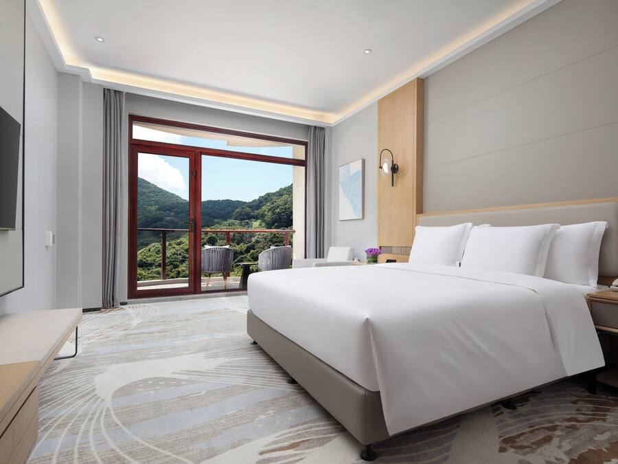 Ein Hotelzimmer mit einem großen Bett, einem Nachttisch mit Telefon, einem Flachbildfernseher, einem Fenster mit Blick ins Freie und einer Schiebetür, die auf einen Balkon führt.