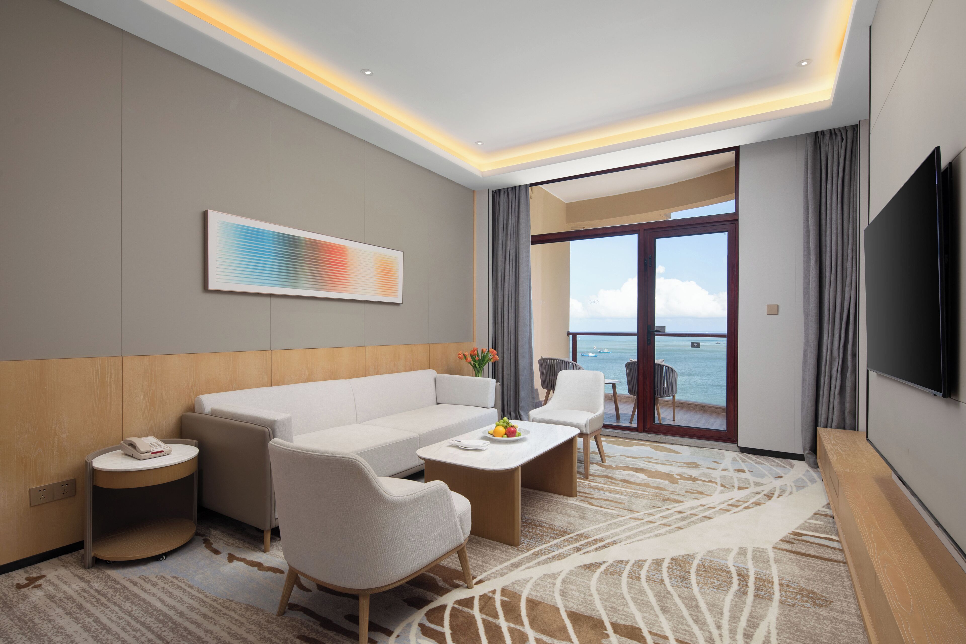 A modern hotel room with a sofa, chairs, a small table, a TV, and a view of the sea through a glass door.