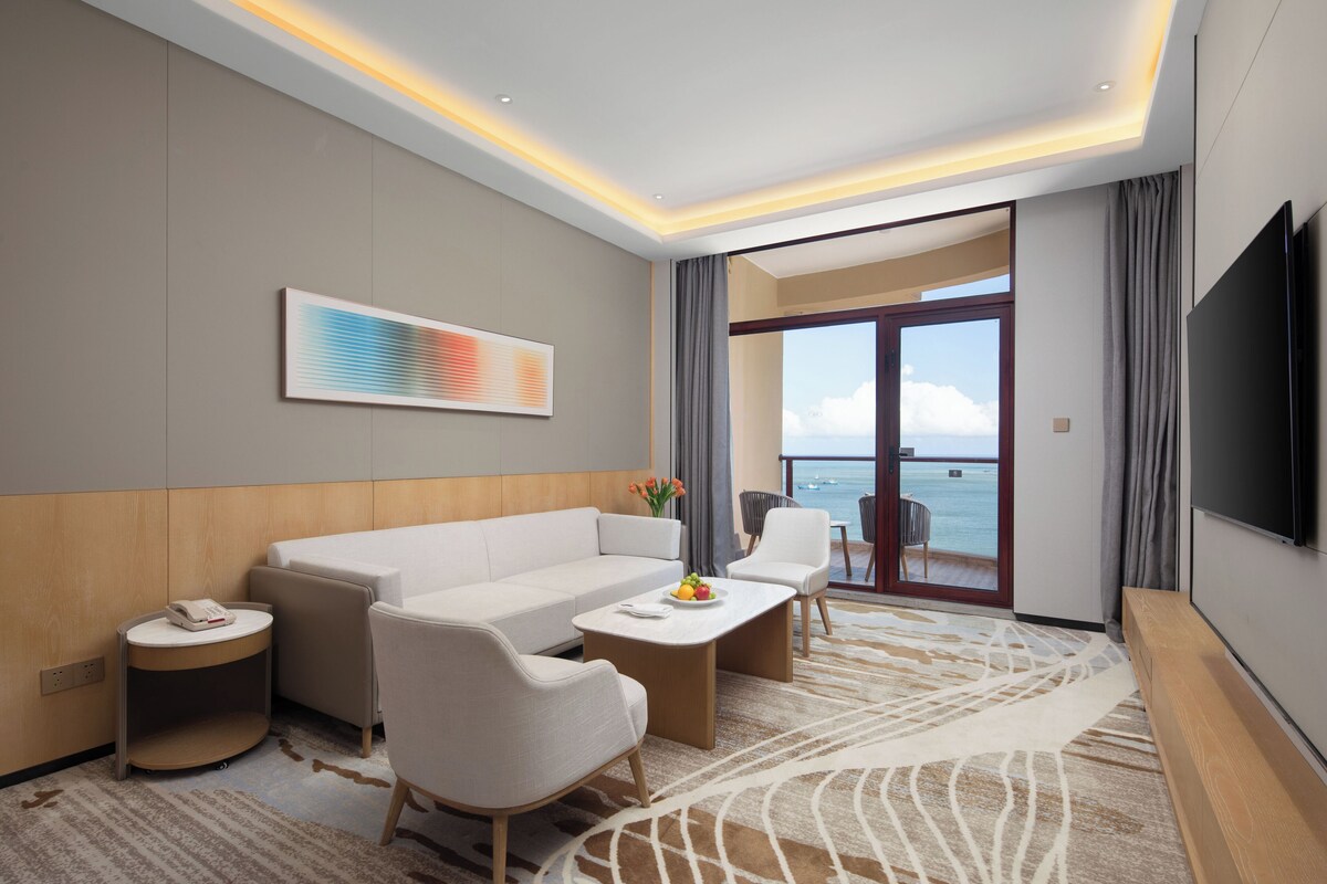 A modern hotel room with a sofa, chairs, a small table, a TV, and a view of the sea through a glass door.