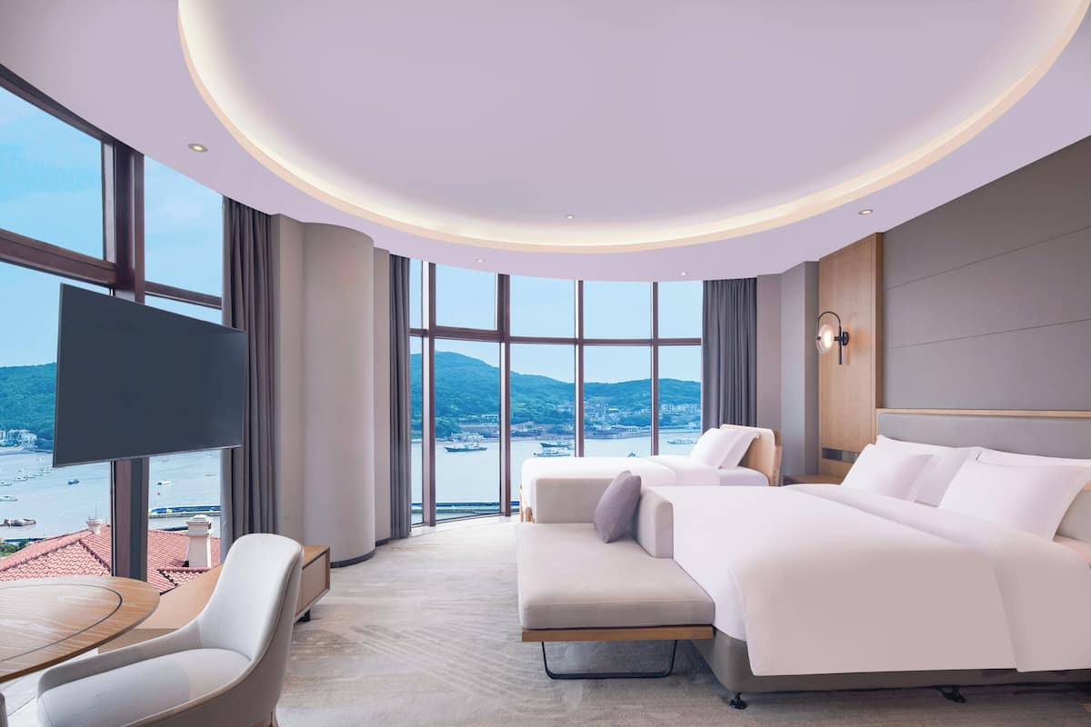 A modern hotel room with a large bed, a sofa, a TV, and a view of the sea.