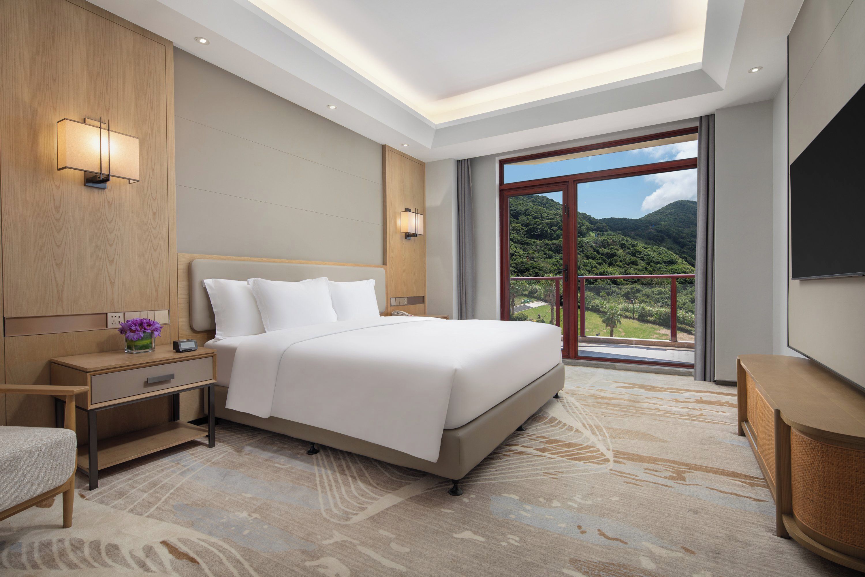 A hotel room with a large bed, bedside tables, a nightstand, a chair, a TV, and a window with a view of a mountainous landscape.