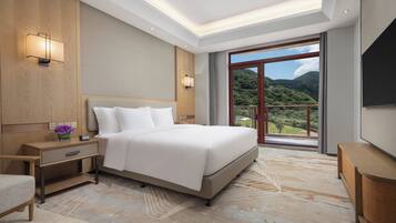 A hotel room with a large bed, bedside tables, a nightstand, a chair, a TV, and a window with a view of a mountainous landscape.