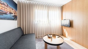 A modern hotel room with a grey sofa, a round wooden table, a TV mounted on the wall, and a large window with sheer curtains. - GuestReady - Tigerlily (Porto)
