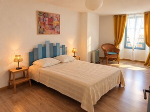 Ein Schlafzimmer mit einem Bett, zwei Nachttischlampen, einem Korbstuhl, einem Heizkörper und einem Fenster mit Vorhängen.