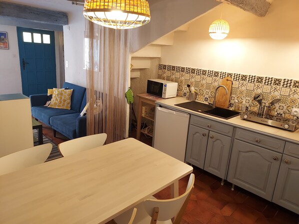 A compact kitchen with a microwave, stove, and sink. There's a dining table with chairs, a blue sofa, and a curtain.