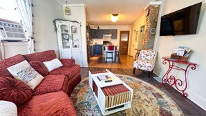 A cozy living room with a red sofa, a coffee table, and a patterned rug. A kitchen and dining area are visible in the background. - Charming Centennial Cottage-Downtown! (Rapid City)