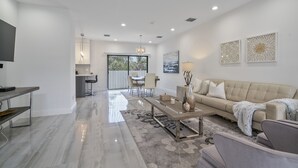 A modern living room with a sofa, coffee table, and a TV. There is a dining area with chairs and a table. The room has a large window and a view of the outdoors. - New Townhome near Wilton Manors/Las Olas (Fort Lauderdale)