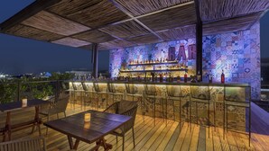 Uma área de bar ao ar livre com um deck de madeira, banquetas e uma parede revestida com um letreiro 'BAR'.