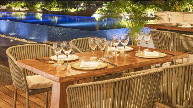 Uma área de refeições com mesa de madeira posta para seis pessoas, rodeada por cadeiras de vime, com vista para uma piscina e um elemento aquático iluminado em azul.