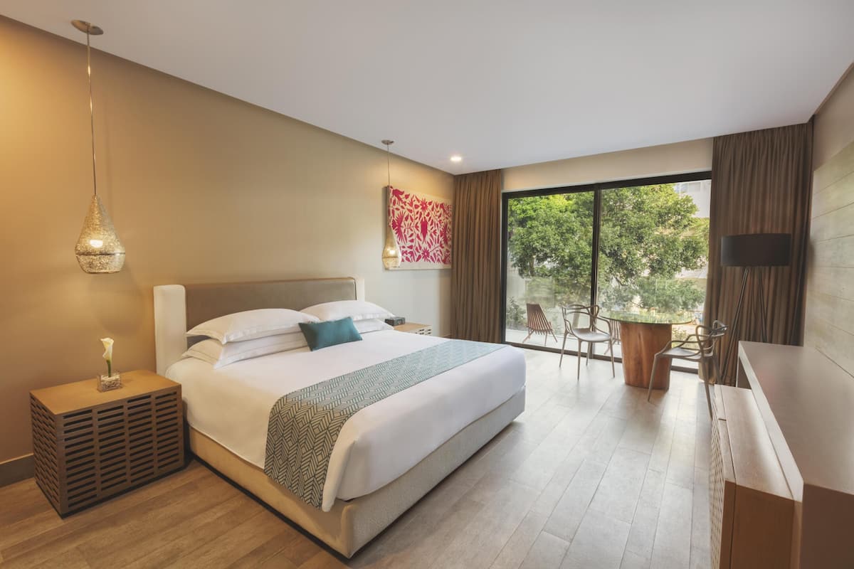 A modern hotel room with a large bed, a desk, and a balcony with a view of trees.