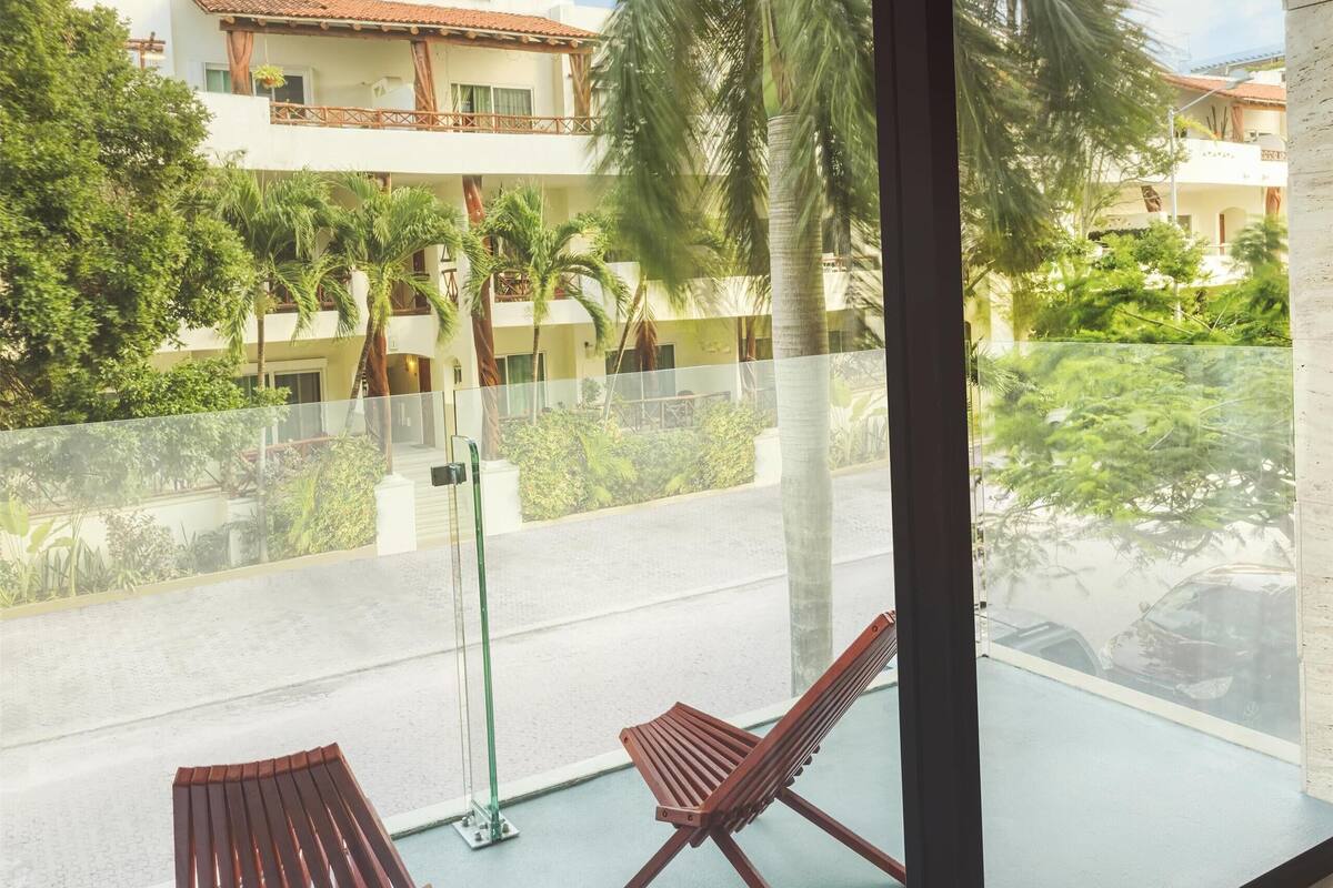 A balcony with two wooden chairs and a glass railing, overlooking a residential area with palm trees and houses.
