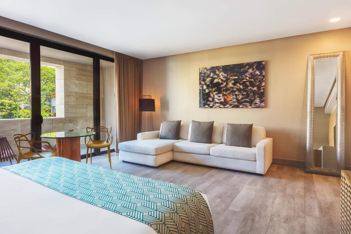 A modern hotel room with a large bed, a glass table, two chairs, a sofa, and a large window offering a view of greenery.