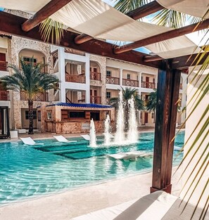 A hotel pool area with a fountain, palm trees, and a multi-story building with balconies.