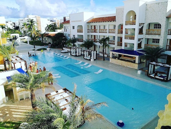 A resort pool area with multiple levels of buildings, palm trees, and lounge chairs.