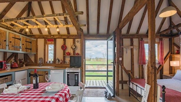 A rustic room with a wooden ceiling, a dining table with a checkered tablecloth, a bed with a red blanket, and a view of the outdoors. - The Granary by Bloom Stays - cute lodge sleeps 2 guests  in 1 bedroom (Canterbury)